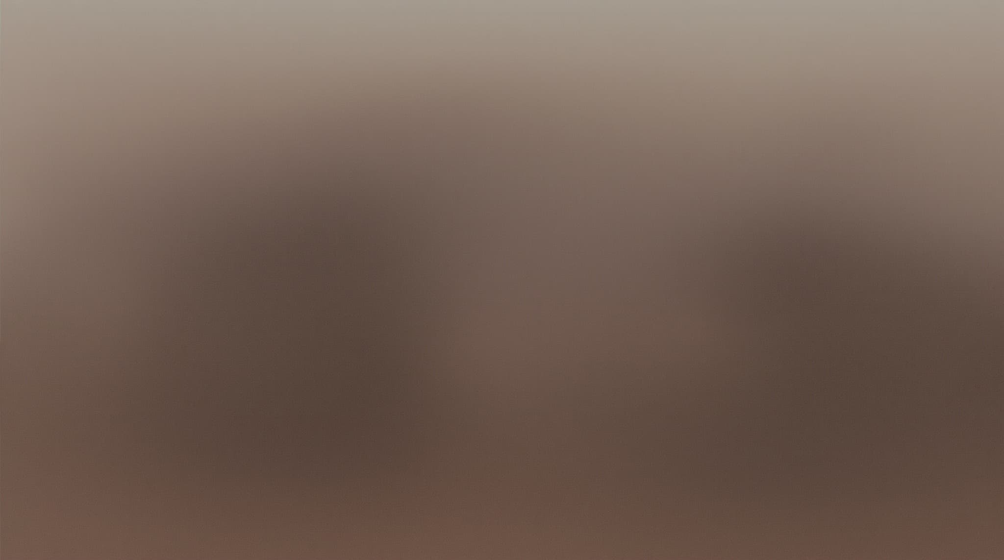 A couple seen from behind on a sofa, each holding a phone showing a parent photo, with a laptop in front previewing a soft pastel AI baby image in warm afternoon light.
